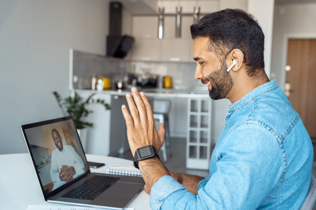 Profile portrait shot of young indian man sitting at home desk front of laptop greeting prof on screen. Remote student practice foreign language communicate online with native speaker using internet